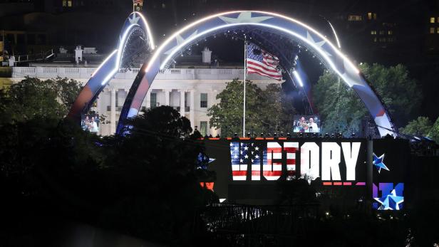 Große Bühne mit leuchtendem Schriftzug „VICTORY“, US-Flagge und dem Weißen Haus im Hintergrund bei Nacht.