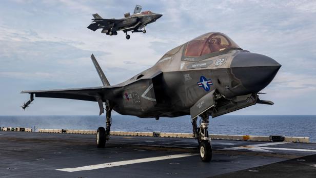 Zwei US-Kampfjets vom Typ F-35 befinden sich auf einem Flugzeugträger, einer steht auf dem Deck, der andere landet gerade.