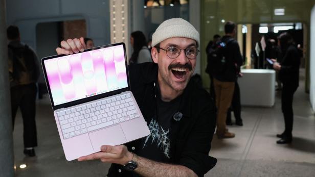 Ein Mann mit Mütze und Brille hält ein rosa Laptop hoch und grinst begeistert in einem Ausstellungsraum.