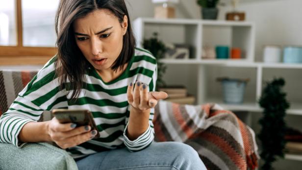 Eine Frau schaut irritiert auf ihr Smartphone und gestikuliert fragend auf dem Sofa.