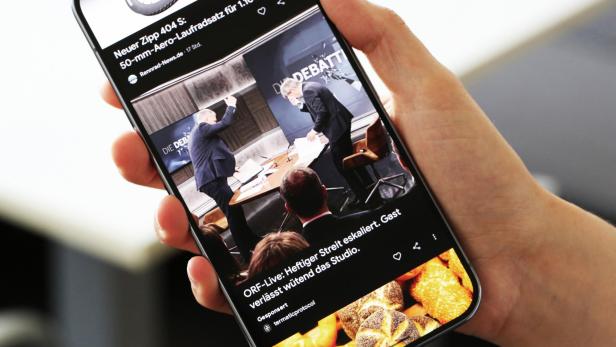 Eine Hand hält ein Smartphone, das einen eskalierenden Streit in einer ORF-Live-Debatte zeigt, bei der ein Gast wütend das Studio verlässt.