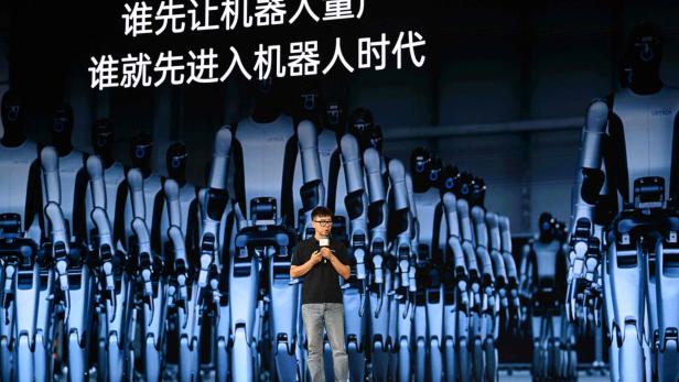 Vortragender vor riesiger Robotergruppe mit chinesischer Schrift auf der Leinwand.