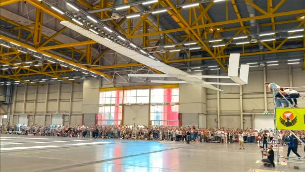 Ein riesiger Papierflieger wird in einer Halle mit Publikum gestartet.