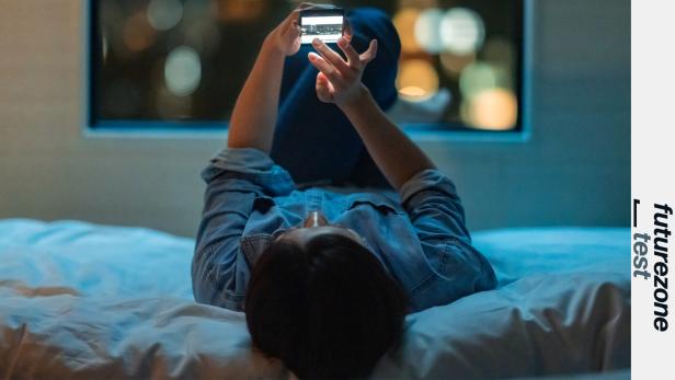 Person liegt auf dem Bett und hält ein Smartphone hoch.