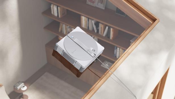 Ein ECOVACS Winbot W3 Omni Fensterputzroboter reinigt eine Glasfläche über einem Bücherregal.