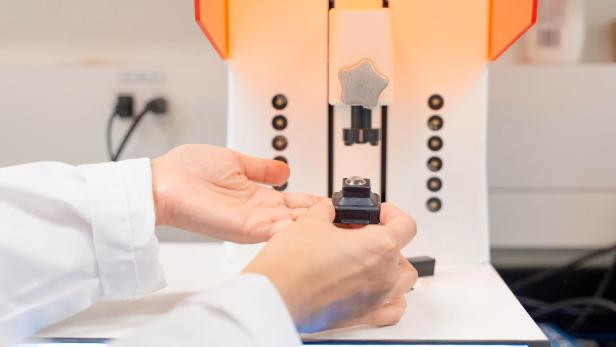 Hände halten eine kleine Linse vor einem 3D-Drucker.