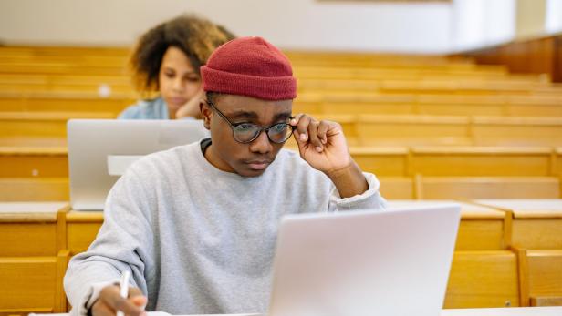 Studierender mit roter Mütze arbeitet in einem Hörsaal am Laptop, während hinter ihm eine Kommilitonin ebenfalls mit Computer sitzt.