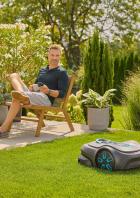 Ein Mann entspannt im Garten, während ein Rasenmähroboter das Gras mäht.