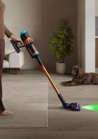 Eine Frau saugt mit einem Dyson-Staubsauger den Boden, während ein Hund zusieht.