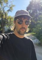 Ein Mann mit Sonnenbrille und Kappe macht ein Selfie im Freien.
