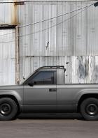 Ein grauer, kantiger Slate-E-Pick-up mit kurzer Ladefläche steht vor einer Wellblechwand.