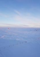 Weite, schneebedeckte Eislandschaft der Antarktis mit kleinen Forschungsstationen und einem Zaun unter einem hellblauen Himmel.