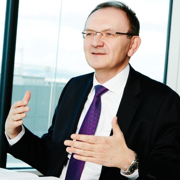 Ein Mann mit Brille und Anzug gestikuliert vor einem Fenster.