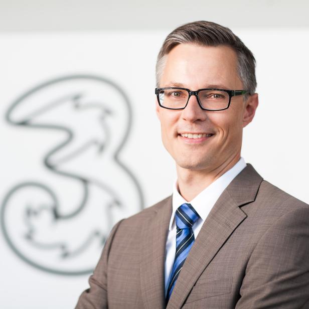 Ein Mann mit Brille und Anzug vor dem Logo von Drei.