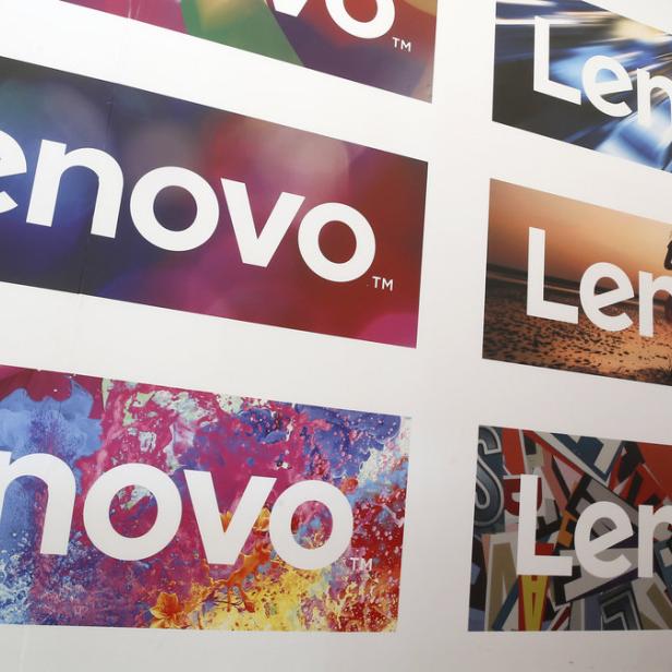 Mehrere Lenovo-Logos mit verschiedenen Hintergründen sind an einer Wand angebracht. Ein Mann sitzt davor und benutzt einen Laptop.