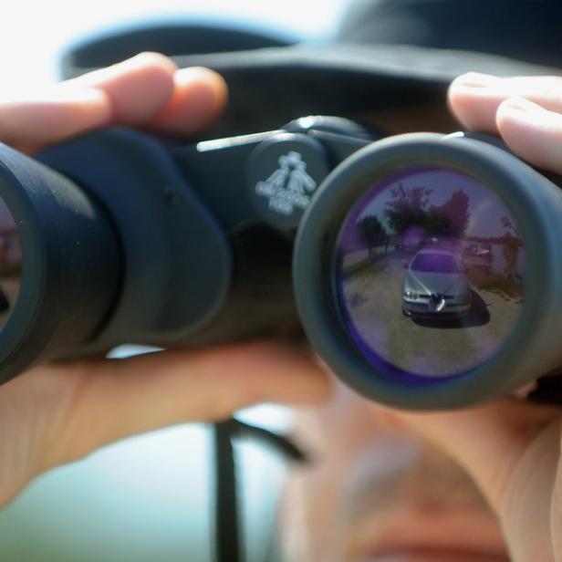 Eine Person blickt durch ein Fernglas, in dem ein silbernes Auto zu sehen ist.