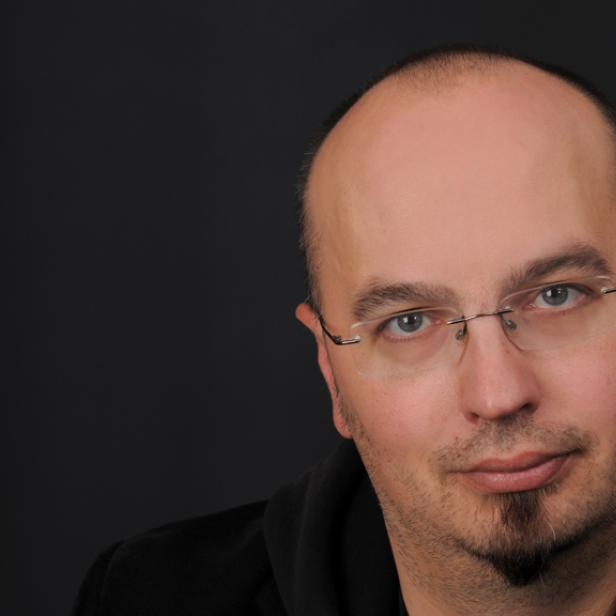 Porträt eines Mannes mit Brille und Bart vor einem dunklen Hintergrund.