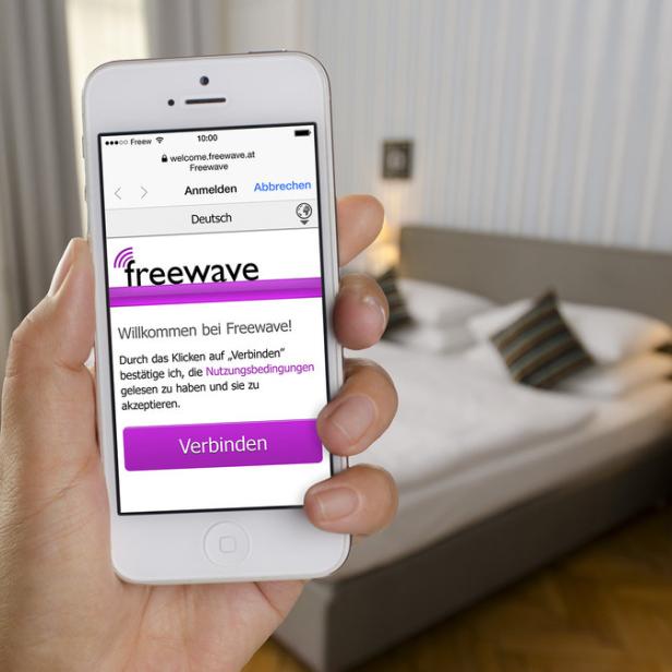 Eine Hand hält ein Smartphone mit der Freewave-Anmeldeseite in einem Hotelzimmer.