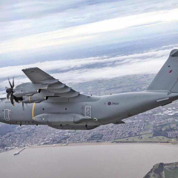 Ein graues Militärflugzeug vom Typ Airbus A400M fliegt über eine Küstenstadt.
