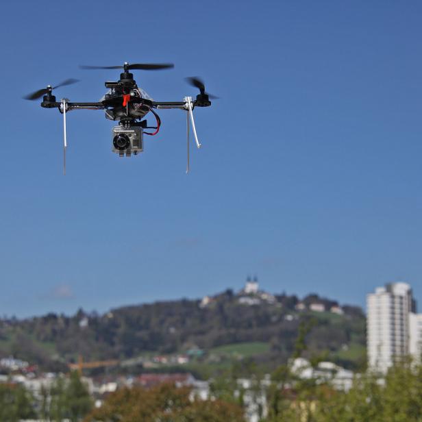 Eine Drohne mit Kamera fliegt vor blauem Himmel über eine Stadt.