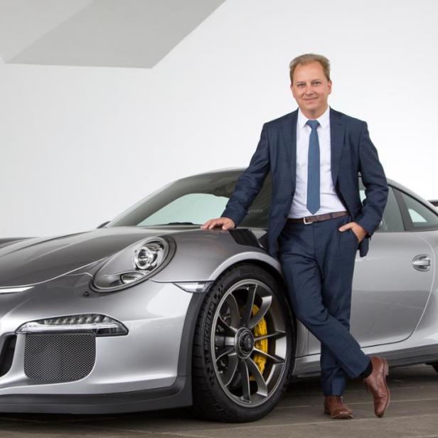 Ein Mann im Anzug posiert neben einem silbernen Porsche mit dem Kennzeichen S GO 5015.