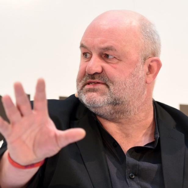 Ein Mann mit Bart gestikuliert vor einem weißen Hintergrund mit dem Logo einer Organisation.