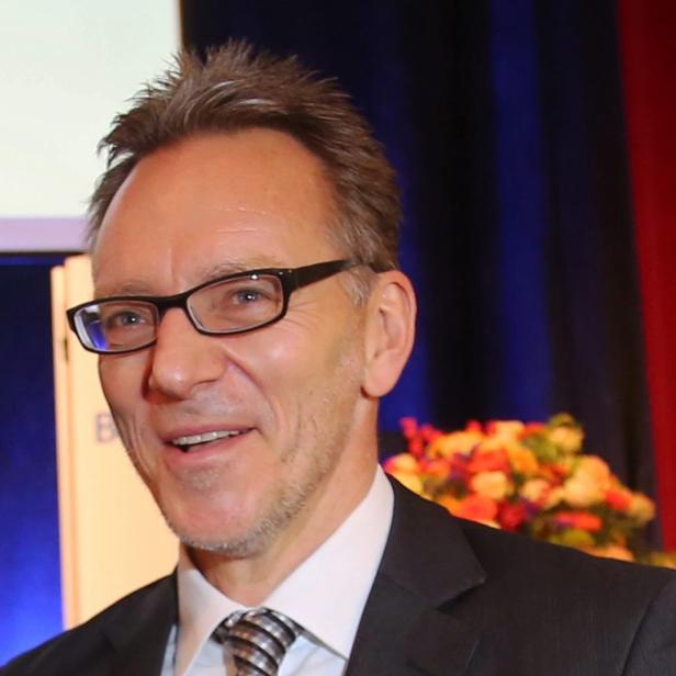 Ein lächelnder Mann mit Brille und Anzug vor einem roten Vorhang.