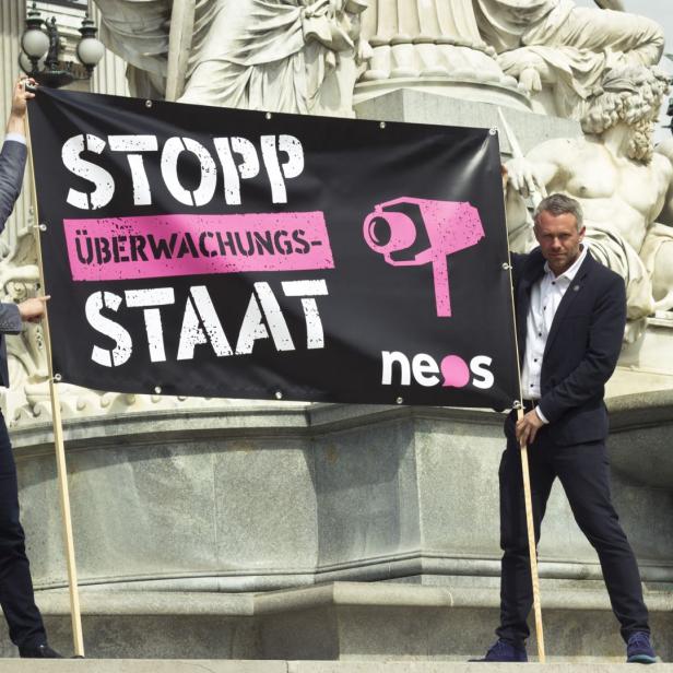 Zwei Männer halten ein Banner mit der Aufschrift „Stopp Überwachungs-Staat“ vor einem Gebäude.