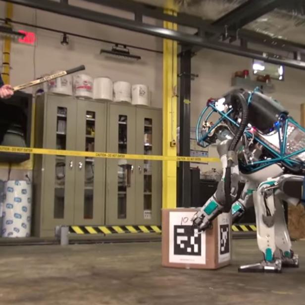 Ein Mann mit einem Hockeyschläger beobachtet, wie ein humanoider Roboter eine Kiste bewegt.
