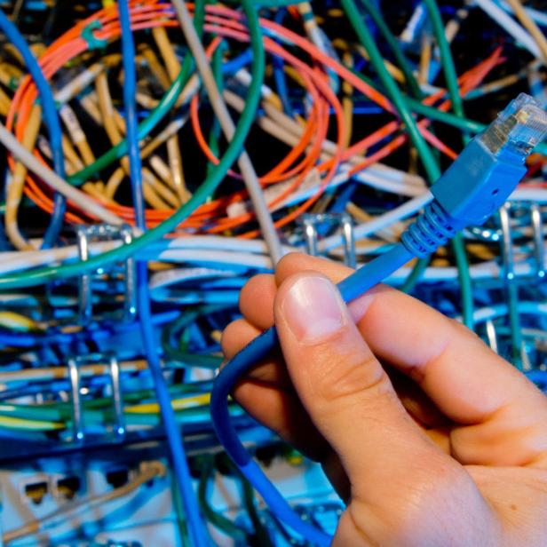 Eine Hand hält ein blaues Ethernet-Kabel vor einem Kabelsalat.