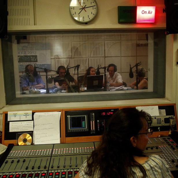 In einem Radiostudio arbeitet eine Frau am Mischpult, während eine Gruppe Personen in einem Aufnahmeraum zu sehen ist.