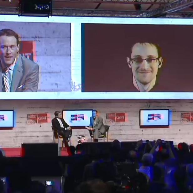 Zwei Männer werden auf einer Konferenz auf Bildschirmen gezeigt, darunter Edward Snowden.