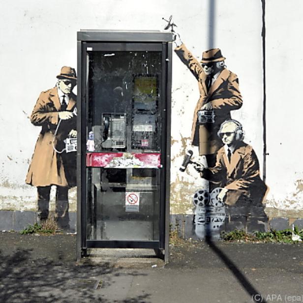 Ein Banksy-Graffiti zeigt drei Männer in Trenchcoats, die eine Telefonzelle abhören.