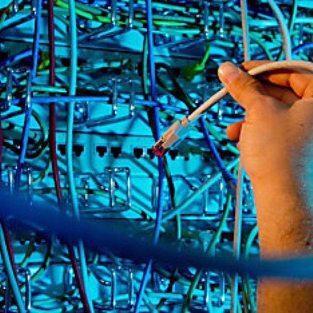 Eine Hand steckt ein Netzwerkkabel in einen Serverraum mit vielen Kabeln.