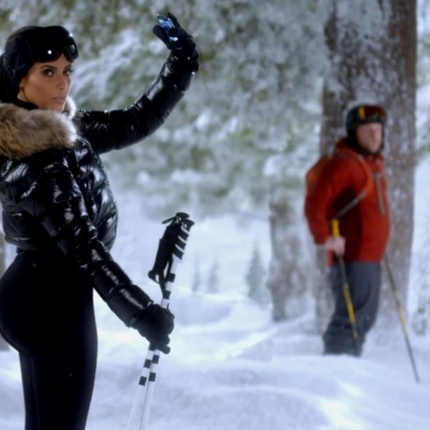 Eine Frau in Skikleidung macht ein Selfie im Schnee, während zwei Männer im Hintergrund stehen.