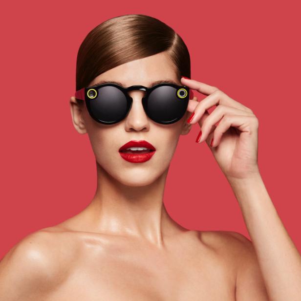 Eine Frau mit roten Lippen trägt eine schwarze Sonnenbrille vor einem roten Hintergrund.