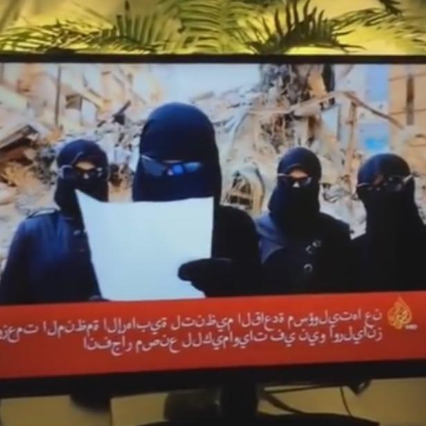 Auf einem Fernsehbildschirm sind vier verschleierte Frauen vor einem zerstörten Gebäude zu sehen.