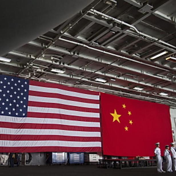 Die Flaggen der USA und Chinas hängen in einer Halle, während Matrosen in Weiß Spalier stehen.