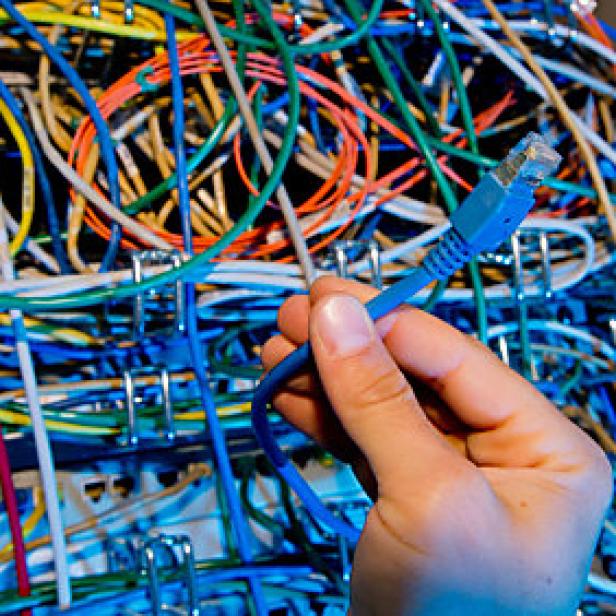 Eine Hand hält ein blaues Netzwerkkabel vor einem Gewirr von bunten Kabeln.