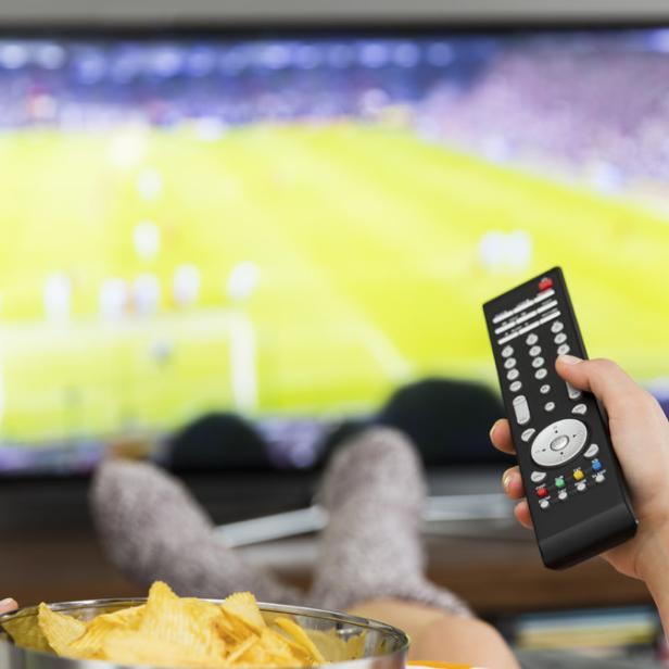 Eine Person entspannt sich mit Chips und Fernbedienung vor dem Fernseher, auf dem ein Fußballspiel läuft.