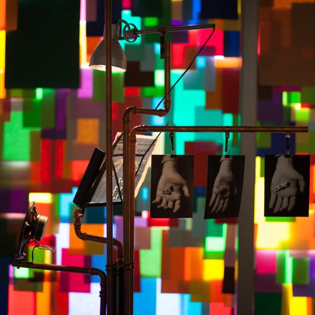 Eine Installation mit einer Lampe, Lautsprechern und Fotos von Händen vor einem farbenfrohen Hintergrund.