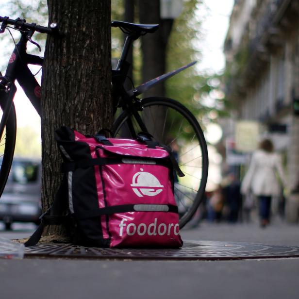 Ein Fahrrad und eine rosafarbene Foodora-Liefertasche lehnen an einem Baum in einer Stadt.