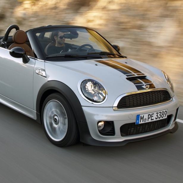 Ein silberner Mini Cooper Roadster fährt auf einer asphaltierten Straße.