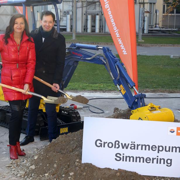 Zwei Personen halten Spaten vor einem Bagger und einem Schild „Großwärmepumpe Simmering“.