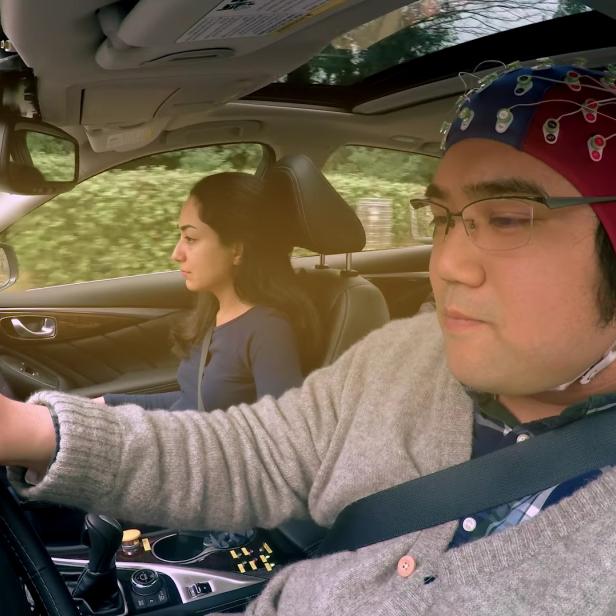 Ein Mann mit einer EEG-Haube fährt ein Auto, eine Frau sitzt auf dem Beifahrersitz.