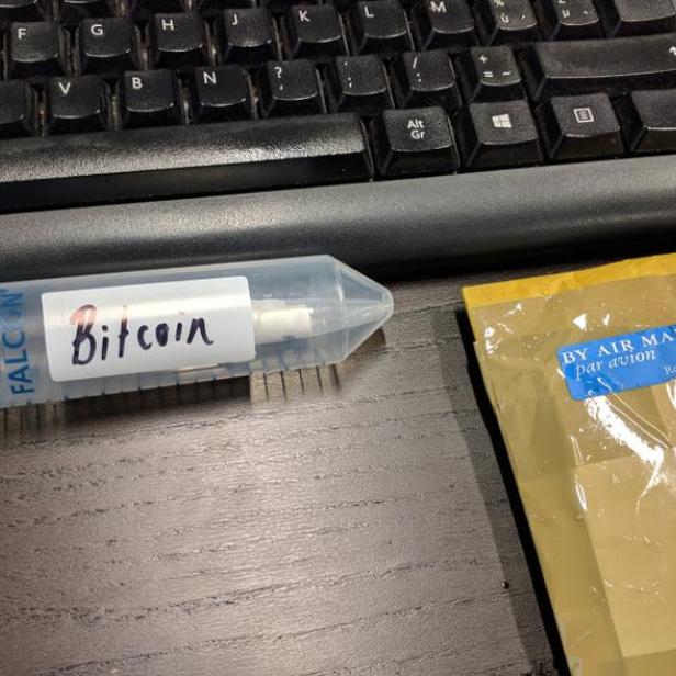Eine Falcon-Tube mit der Aufschrift „Bitcoin“ liegt neben einem Briefumschlag auf einem Schreibtisch.