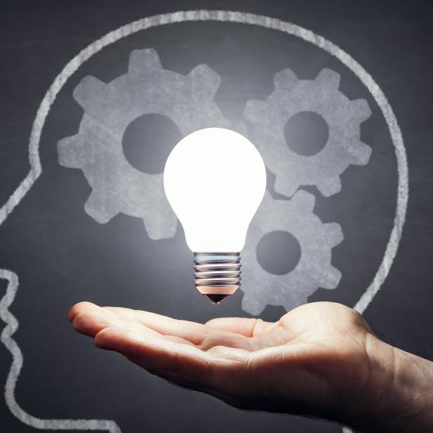 Eine Hand hält eine leuchtende Glühbirne vor einer Tafel mit einem Kopf und Zahnrädern.