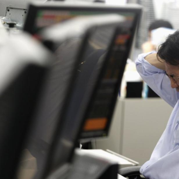 Ein Mann sitzt frustriert vor mehreren Computermonitoren und hält die Hände an den Hinterkopf.