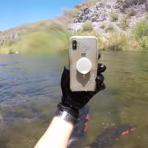 Eine Hand mit Handschuh hält ein nasses iPhone in einem Fluss.