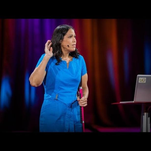 Eine Frau in einem blauen Kleid hält einen Stock in der Hand und spricht auf einer Bühne.